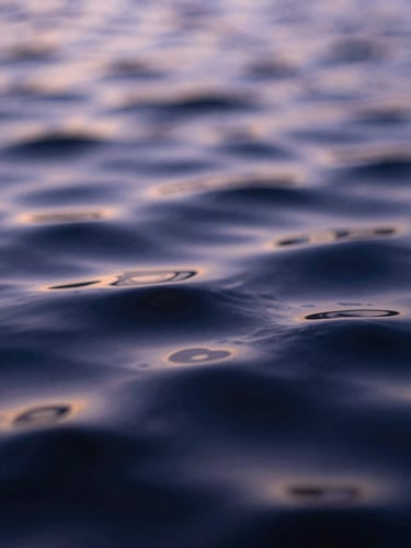 Extreme macro close-up of a textured liquid surface, dark indigo ripples reflecting pale lavender twilight, soft out-of-focus light.