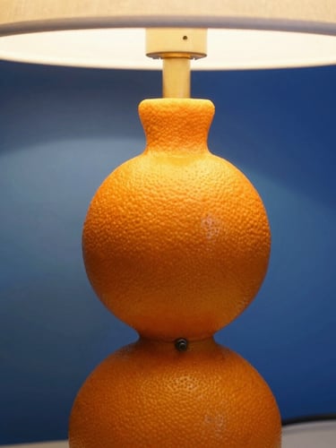 Close-up of a vibrant tangerine sculptural table lamp casting a sharp geometric shadow on a cobalt blue backdrop, warm ambient glow, 35mm lens