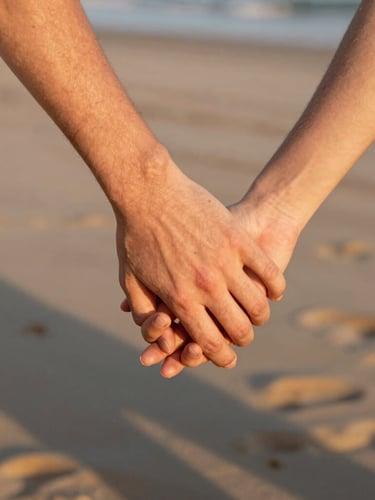 Close-up of two hands gently holding each other on a windy beach, warm golden-hour light casting soft shadows, intimate depth of field, 35mm