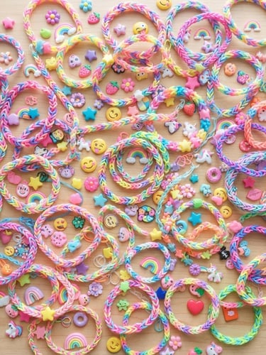 Overhead flatlay of colorful hand-woven rubber band bracelets and 3D puffy stickers scattered on a light wood desk, bright natural daylight, soft shadows