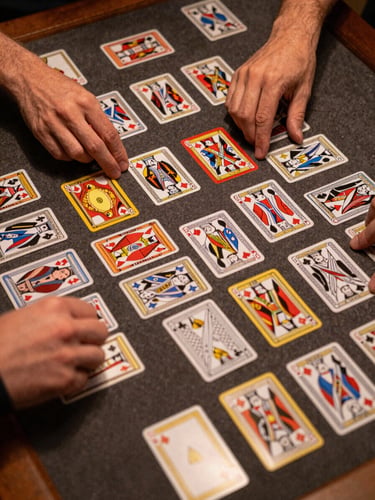 Close overhead shot of trading cards spread across a game table mid-play, two sets of hands reaching in to arrange a play, under honest tungsten indoor light, no faces visible, focus on the cards and the table surface