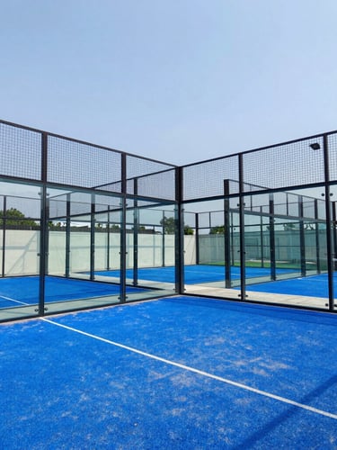 Low-angle architectural photograph of a completed professional padel court with crystal-clear glass walls, vibrant blue turf, under bright Indonesian daylight, structural steel frame visible