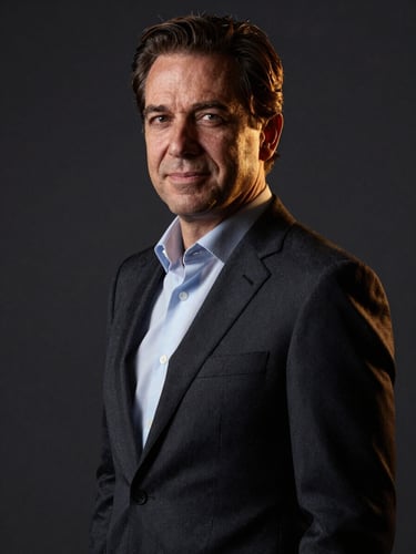 Professional executive portrait of Leo Colella in a dark tailored suit, high-contrast side lighting, deep charcoal background, commanding and warm expression, editorial corporate photography