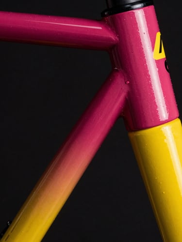 Extreme close-up of a bicycle frame headtube being painted with a deep magenta to electric yellow gradient, fine spray mist catching a sharp spotlight, dark background.