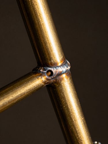 Extreme close-up of a brass fillet weld on a raw steel bicycle frame, warm editorial photography, dramatic studio spotlighting, rich textures, deep shadows, 35mm