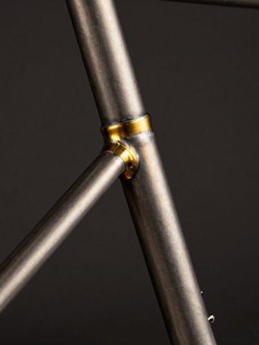 Extreme close-up of a perfectly aligned raw steel bicycle frame joint, glowing brass fillet brazing, dramatic studio spotlighting, deep charcoal background, 35mm.