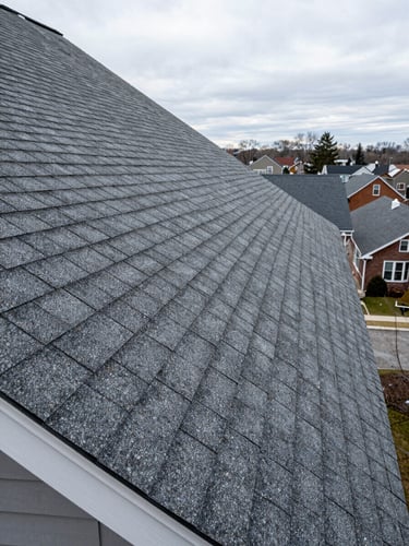 Wide panoramic shot of a steep residential roofline freshly shingled, viewed from ground level looking up to the right, natural overcast daylight, asphalt shingle rows crisp and even, ridge cap tight, fascia and drip edge visible at bottom left, Rochester neighborhood rooftops in background distance