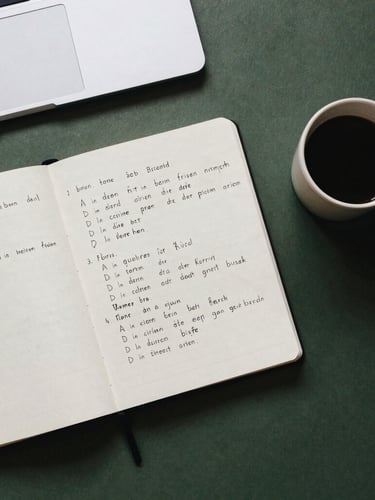 Overhead flat-lay of a clean study desk, an open notebook with handwritten German declension charts, a ceramic coffee cup on the side, soft natural overcast daylight, muted earth tones and deep forest green accents, 35mm film aesthetic.