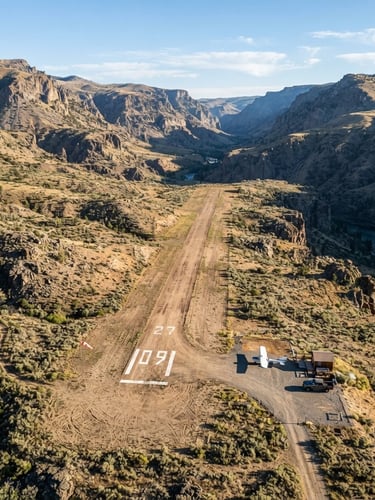 Wide aerial overview of a single dirt runway in a rugged Idaho canyon basin, crisp morning daylight casting sharp shadows across the dry terrain, desaturated earth tones with high-contrast runway markings.