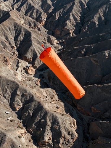 High-contrast aerial photograph of an orange wind sock blowing taut against a rugged Idaho desert canyon background, crisp morning light, desaturated earth tones