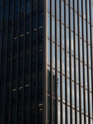 Macro architectural detail of a modern steel and glass skyscraper corner, sharp high-contrast daylight casting dramatic geometric shadows, deep navy tones, minimalist editorial photography