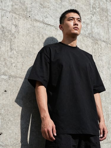 Low-angle heroic shot of a man wearing a heavy-weight black oversized t-shirt, standing against a raw concrete brutalist wall, harsh direct sunlight, high-contrast shadows