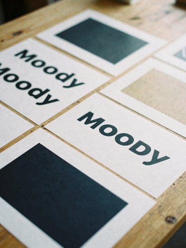Moody close-up 35mm film photograph of printed brand assets and typography proofs laid out on a raw wooden workshop table, overcast natural light, high-contrast shadows