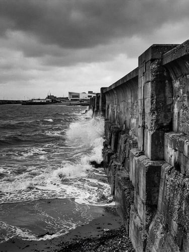 Wide-angle black and white film photograph of the brutalist Margate sea wall on an overcast day, high-contrast dark sea-navy water crashing, raw texture, negative space on the left