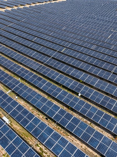 Wide aerial drone shot of massive utility-scale solar arrays stretching to the horizon under harsh midday sun, emphasizing scale and physical reality.