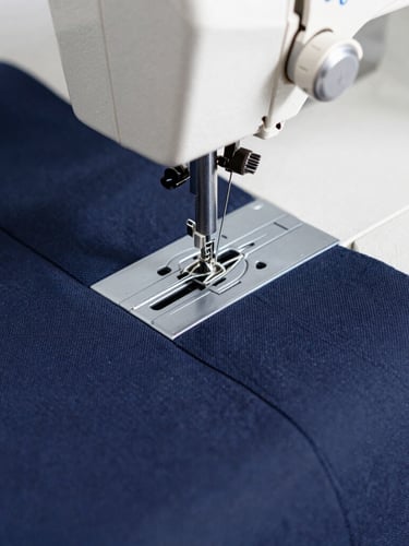 Macro close-up of heavy-duty sewing machine stitching a navy blue corporate jacket, bright natural daylight, sharp focus on textile weave, 35mm photography