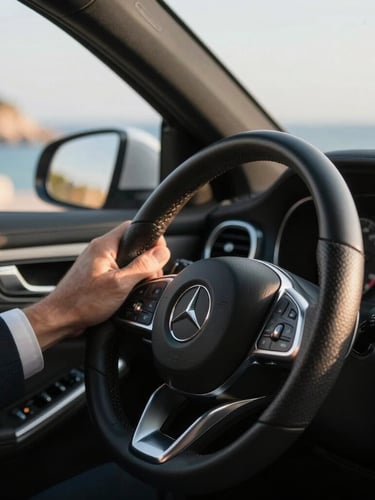 Close-up of a professional driver's neatly dressed hand on the leather steering wheel of a Mercedes-Benz, warm Mediterranean sunlight streaming through the window, soft focus coastal background