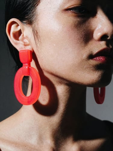 Extreme close-up of a model wearing massive neon-red acrylic earrings, high-contrast studio strobe lighting, sharp black shadows, avant-garde fashion zine style.