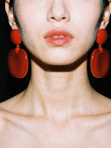 Extreme close-up of a model with a sharp jawline wearing massive, chunky hot-red acrylic earrings that graze her shoulders, harsh direct studio strobe lighting casting deep shadows on a solid black background, 35mm film texture.