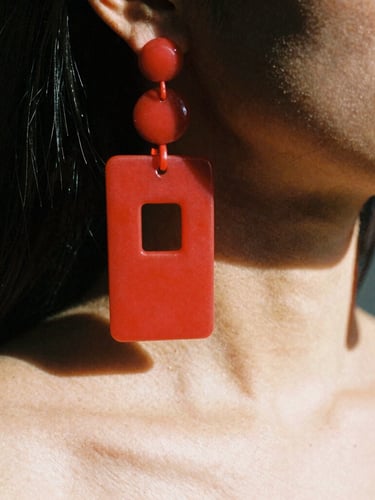 Extreme macro shot of heavy red acrylic earrings hanging to the shoulder of a model, high-contrast direct sunlight, sharp dark shadows, avant-garde fashion photography, 35mm film.