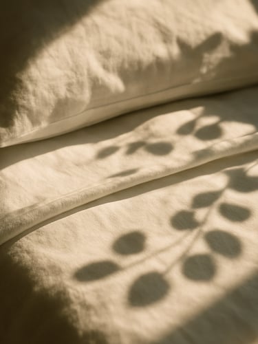 Tight close-up of crisp, perfectly folded white linen sheets on a sunlit bed, warm morning light, shadows of eucalyptus leaves