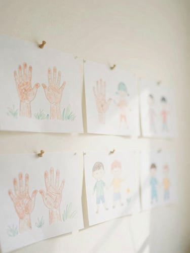 Soft overexposed photograph of children's hand-colored drawings pinned to a sunny cream wall, gentle morning light, cozy and airy atmosphere, 35mm