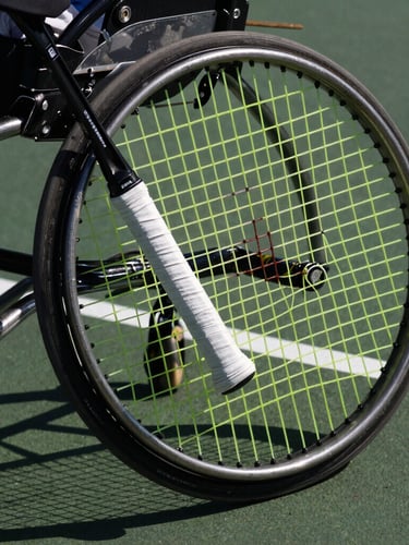 Macro crop of a tennis racket grip and a spinning wheelchair wheel on a dark hard court, intense direct sunlight casting strong shadows, 85mm.