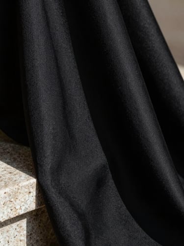 Macro close-up of heavy black silk crepe fabric draped over a stone pedestal, raking natural sunlight highlighting the rich textile weave, deep shadows.