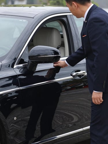 Professional chauffeur in dark uniform, opening the door of a pristine black executive sedan, crisp morning daylight, deep navy and silver tones.