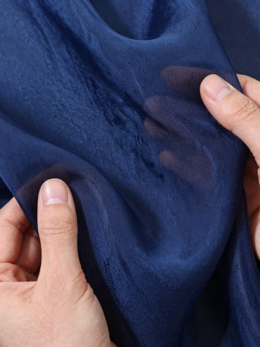 Macro overhead flat-lay of hands holding a premium Turkish organza swatch in deep navy blue, soft side-lit daylight highlighting the delicate weave and translucent texture