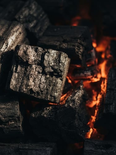 Dramatic side-lit macro shot of high-density Indonesian hardwood charcoal blocks, deep charcoal textures with subtle warm ember-like backlighting, 50mm, industrial premium.
