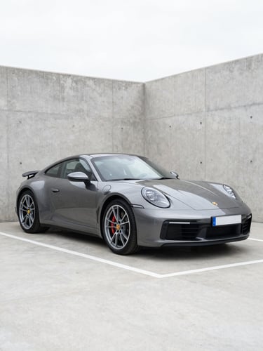 A clean three-quarter shot of a modern dark metallic Porsche 911 parked inside a pristine concrete dealership bay under bright daylight.