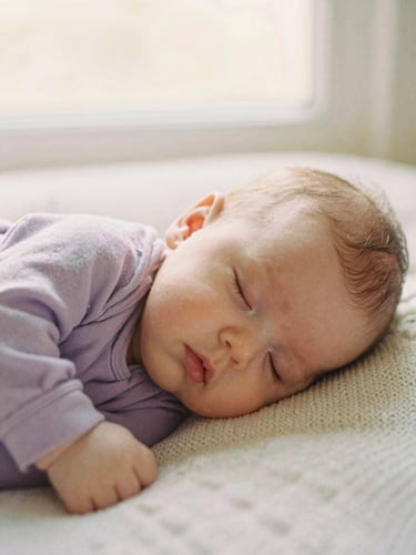 Close-up of a baby sleeping peacefully on a soft knit blanket, warm diffused morning light, gentle lilac and cream tones, editorial style