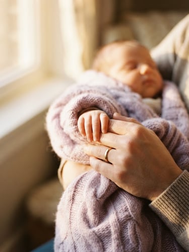 Close-up of a parent gently holding a child's tiny hand wrapped in a soft lilac knitted blanket, warm golden-hour window light, shallow depth of field, 35mm film.