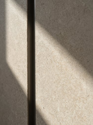 Extreme close-up of aged travertine wall panel meeting a dark steel reveal strip, raking sidelight from the left casting sharp shadows across the stone surface texture, empty architectural interior, no people