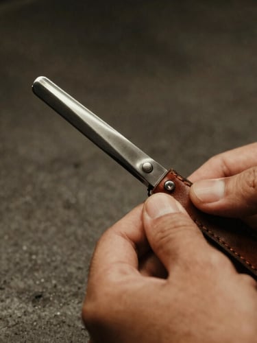 Close-up macro shot of a barber's hands holding a sharp steel razor next to a leather strap, warm side lighting, dark concrete background, rich textures, shallow depth of field.