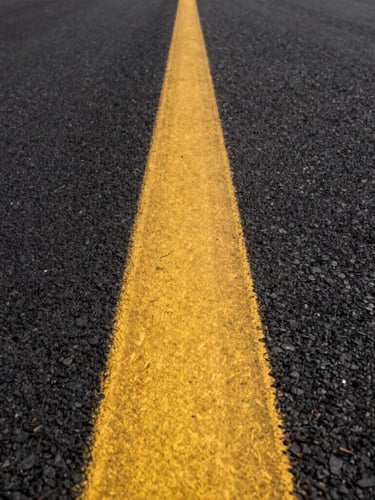 Low-angle perspective shot looking down a long, perfectly straight yellow traffic line on fresh black asphalt, bright morning light casting sharp textures.