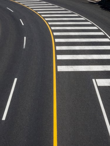 High-angle drone shot of a perfectly striped municipal highway curve under bright midday sun, pristine white and yellow thermoplastic lines stretching into the distance, deep black asphalt texture.