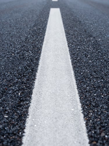 Low-angle perspective shot looking down a perfectly straight, wet white thermoplastic road line on dark asphalt, sharp glass beads reflecting morning light.