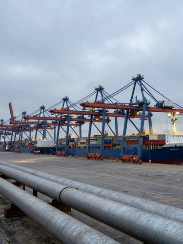 Wide environmental shot of a massive industrial terminal at an overcast harbor, cargo vessels docked, steel pipelines running to the foreground, cool morning light.