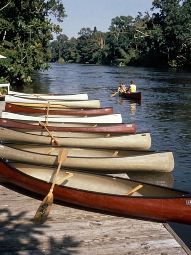 Vintage archival photo of canoes lined up on the wooden docks of the Ipswich River in 1955, warm nostalgic tones, family-owned heritage.