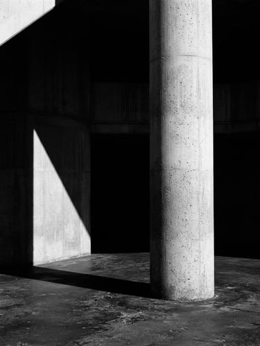 Stark black and white fine art photography, harsh high-contrast directional light, deep architectural shadows, concrete column silhouette, minimalist composition, 35mm film.