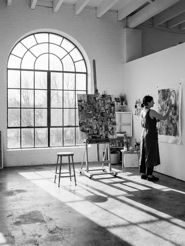 A stark, high-contrast black and white photograph of an art studio with natural light streaming through a large arched window, casting long shadows on clean concrete floors.