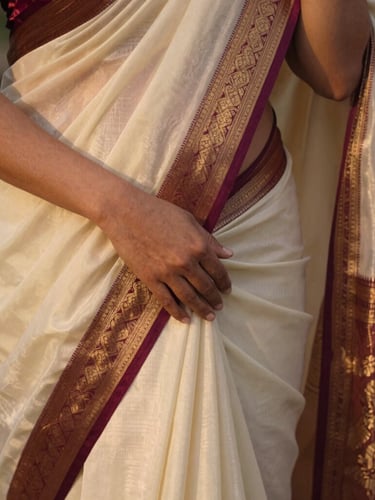 Close-up of a woman's hands gently draping a silk saree, light catching the intricate zari border, soft golden hour light, shallow depth of field, warm editorial photography