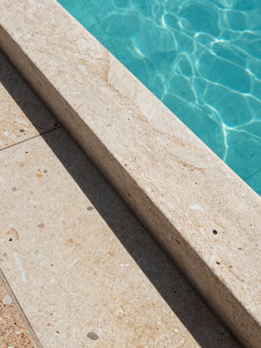 Close-up of natural stone coping and pool edge detail, gresite tile texture visible beneath clear water, Mediterranean midday light casting sharp shadows, documentary framing, no people, architectural precision