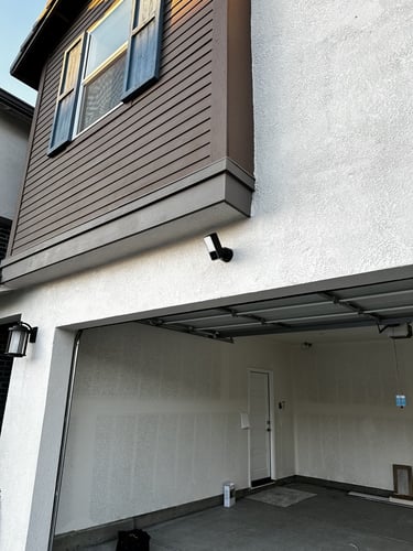 A black outdoor security camera mounted on a white stucco wall above a residential garage door.