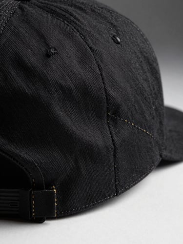 Extreme close-up of a structured black cap on a white studio surface, macro lens revealing raised embroidery thread detail — individual stitches visible, gold thread catching studio strobe light from the upper left, charcoal background at the right edge