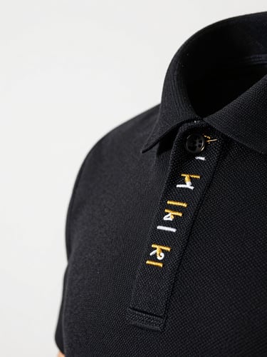 Extreme close-up of premium embroidery on a black polo shirt collar, studio strobe on white background, gold and white thread texture razor-sharp, needle holes visible in fabric weave, subject occupies right two-thirds of frame with generous negative space to the left