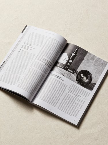 Overhead flatlay of an open editorial magazine spread on a cream linen surface, clean studio light from above, pages showing precise typographic layout and high-contrast black-and-white photography, print quality visible at the fold