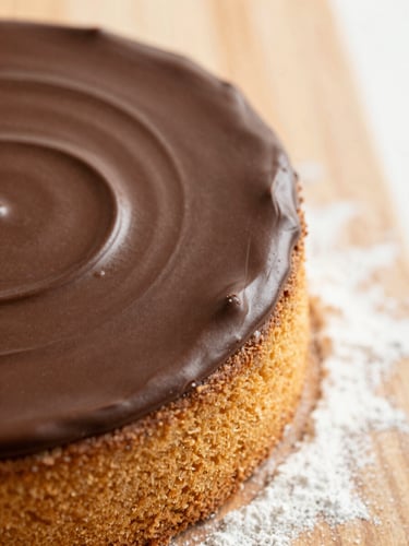 Overhead macro shot of rich chocolate ganache being smoothed over a golden cake layer, dusting of flour on a wooden board nearby, warm light, shallow depth of field